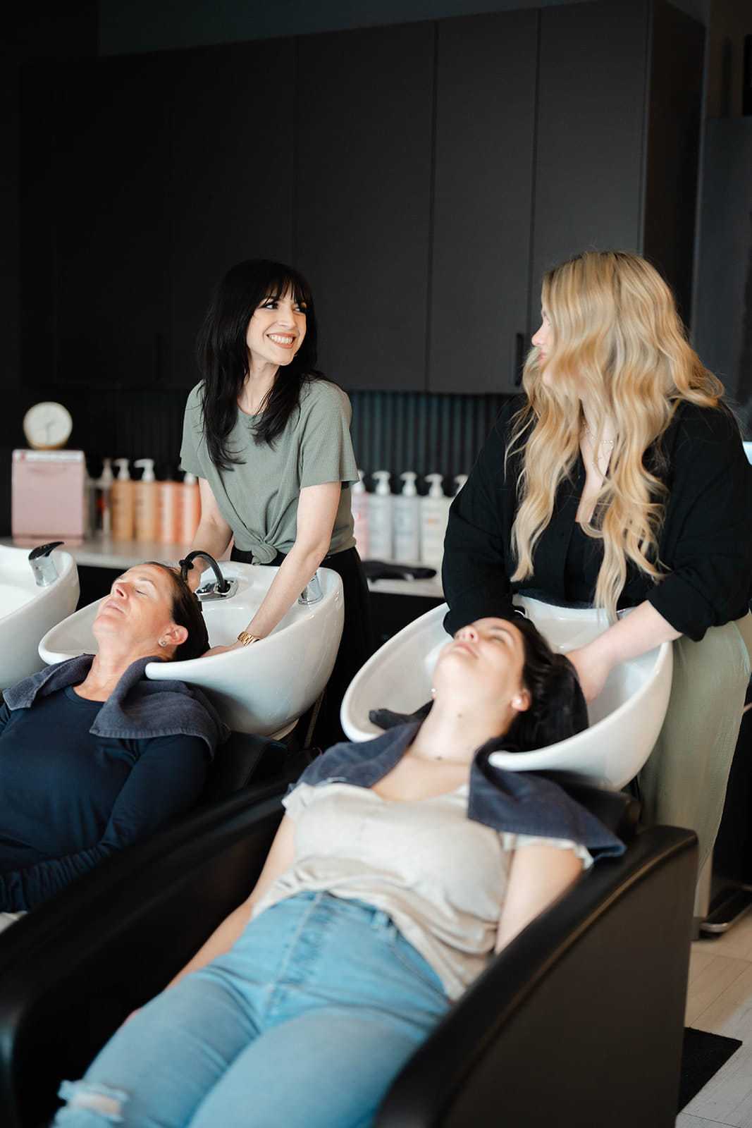 Women getting their hair washed at a modern salon, attended by two stylists.