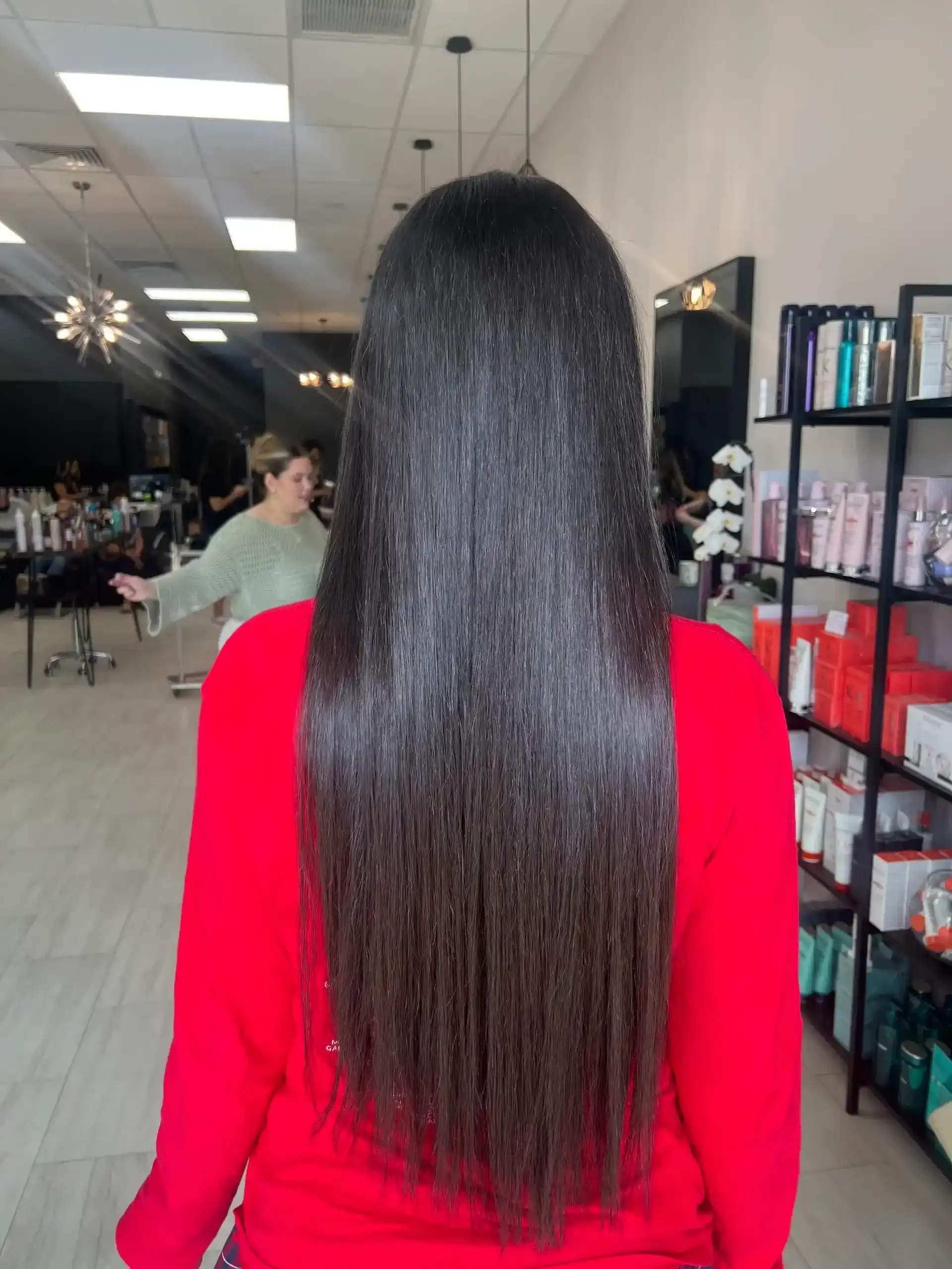 Long, straight hair salon scene with hair products and stylists in the background.