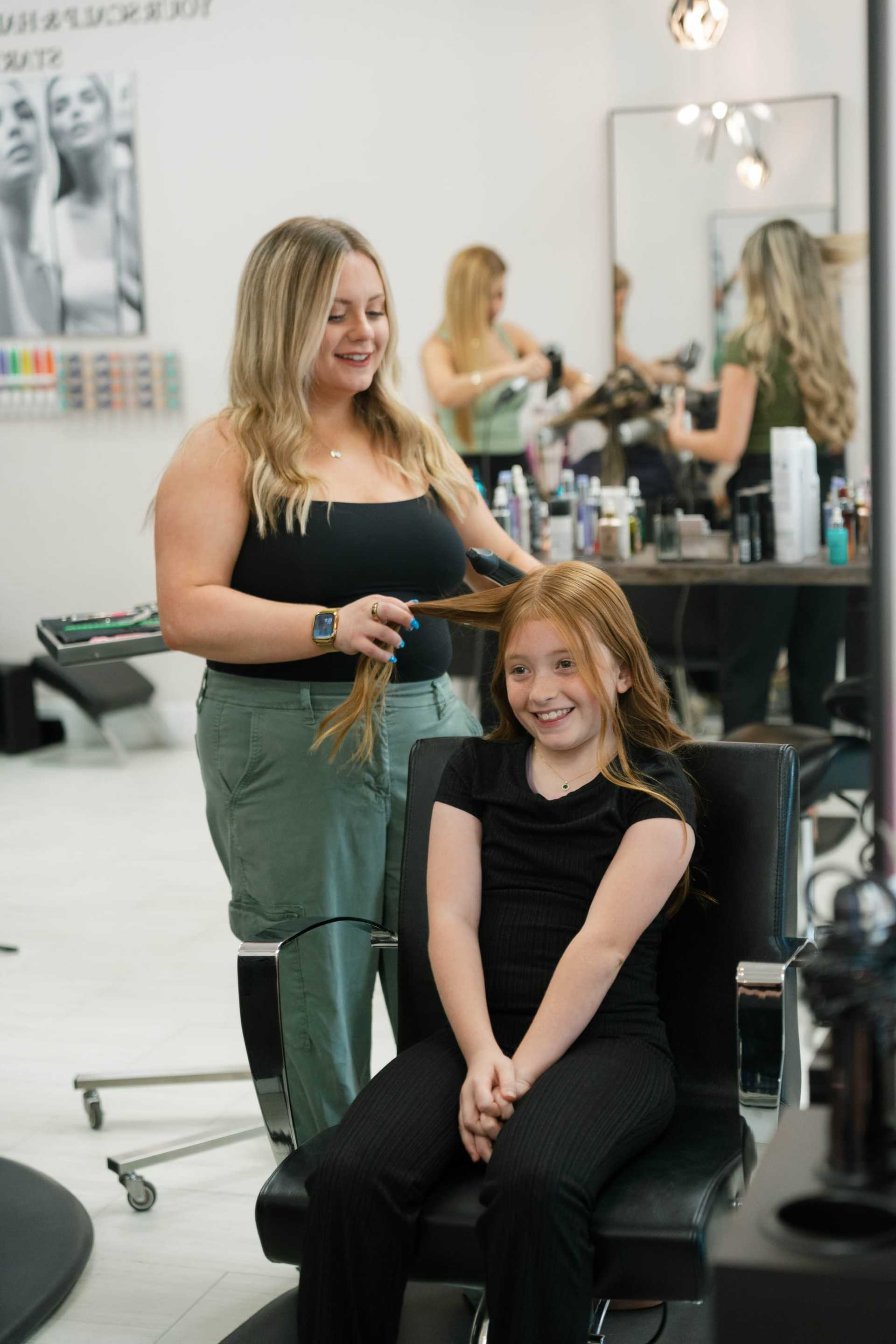 Smiling hairstylist braiding a young girl's hair in a modern salon setting.