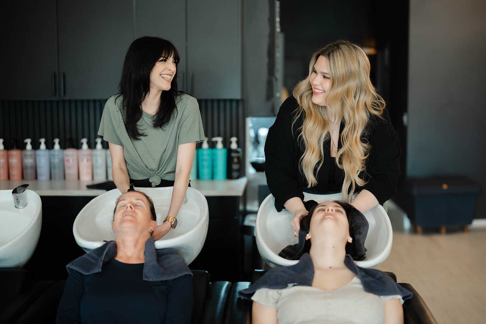 Two hairstylists smiling, washing clients' hair at a salon.