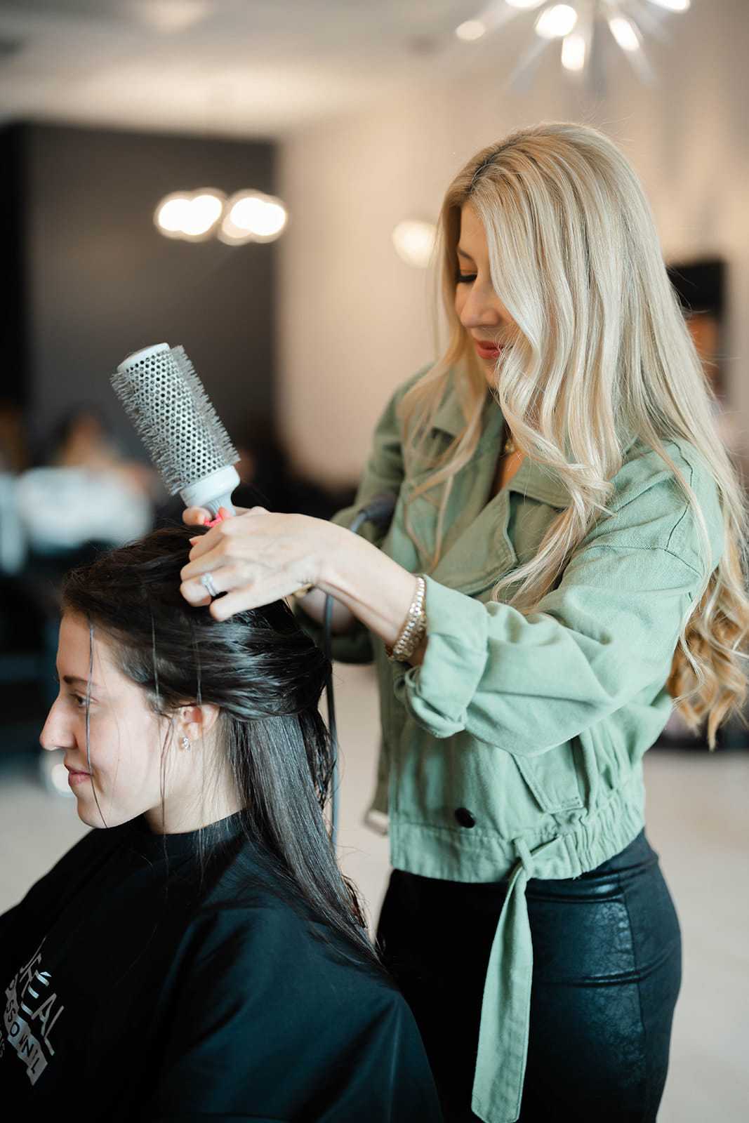 Woman styling another woman's hair in a salon with a round brush.