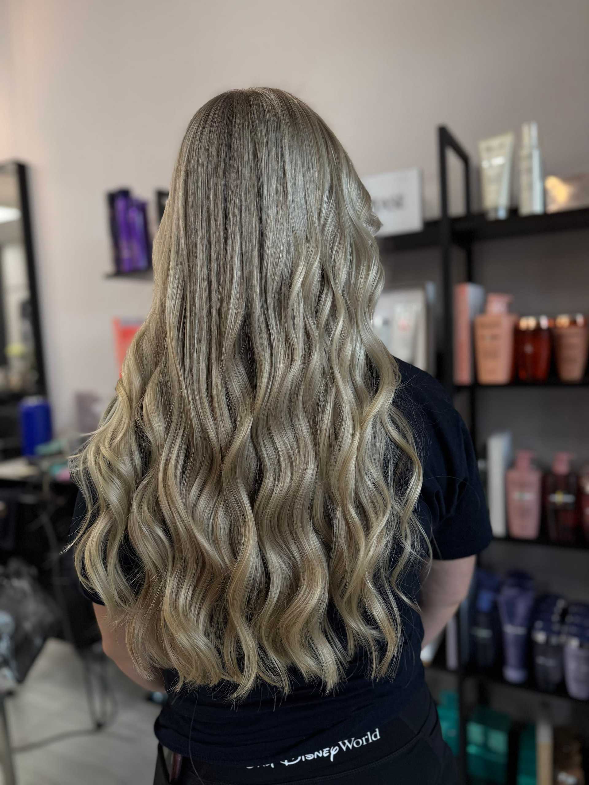 Long wavy blonde hair styled in a salon, with hair products visible in the background.