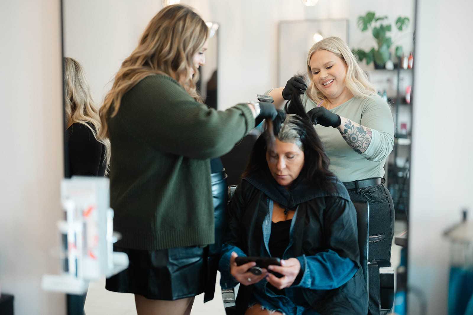 Two stylists dye a woman's hair in a salon as she looks at her phone.
