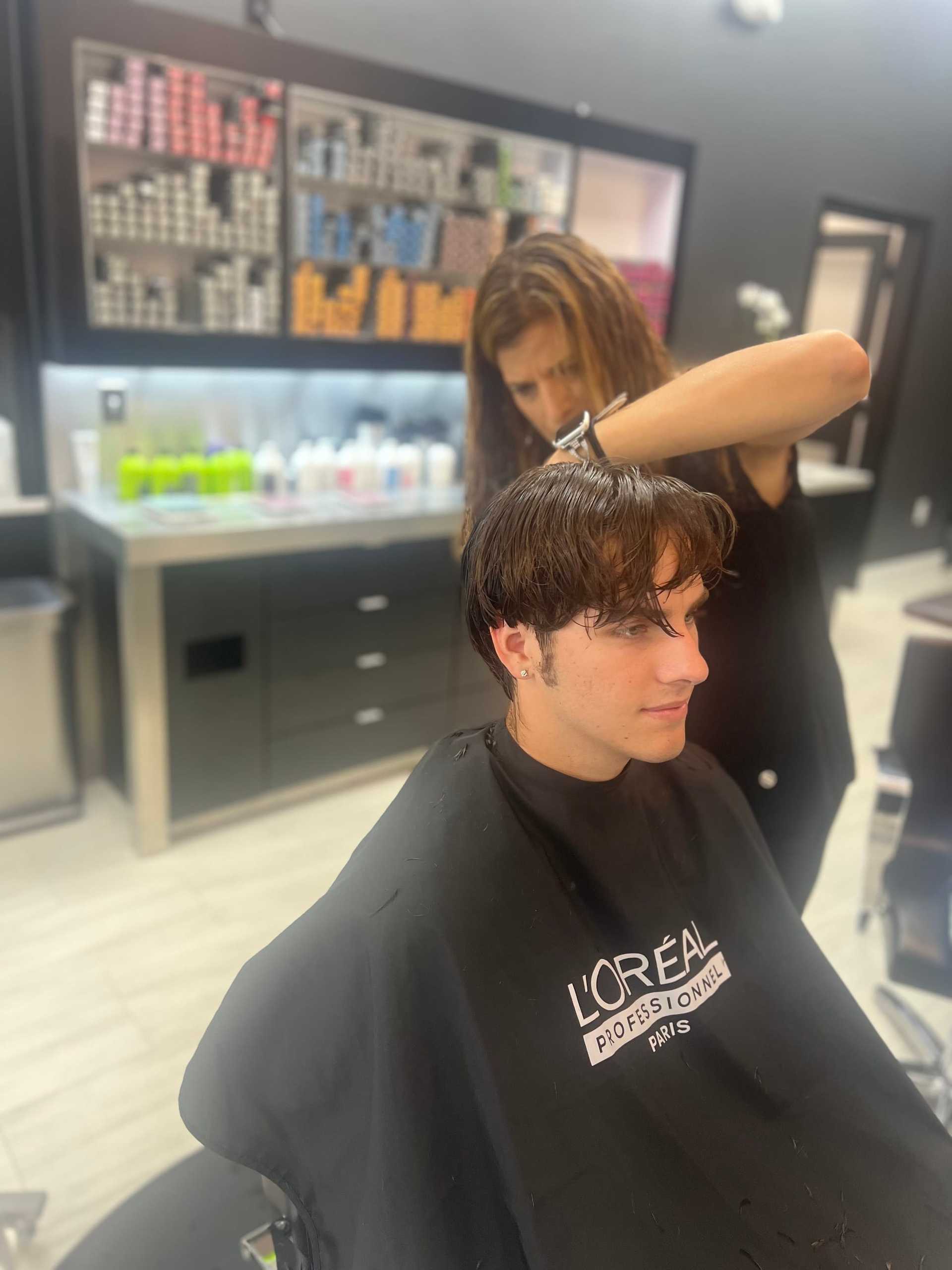 Hairdresser styling a man's hair in a salon. Shelves of hair products in the background.