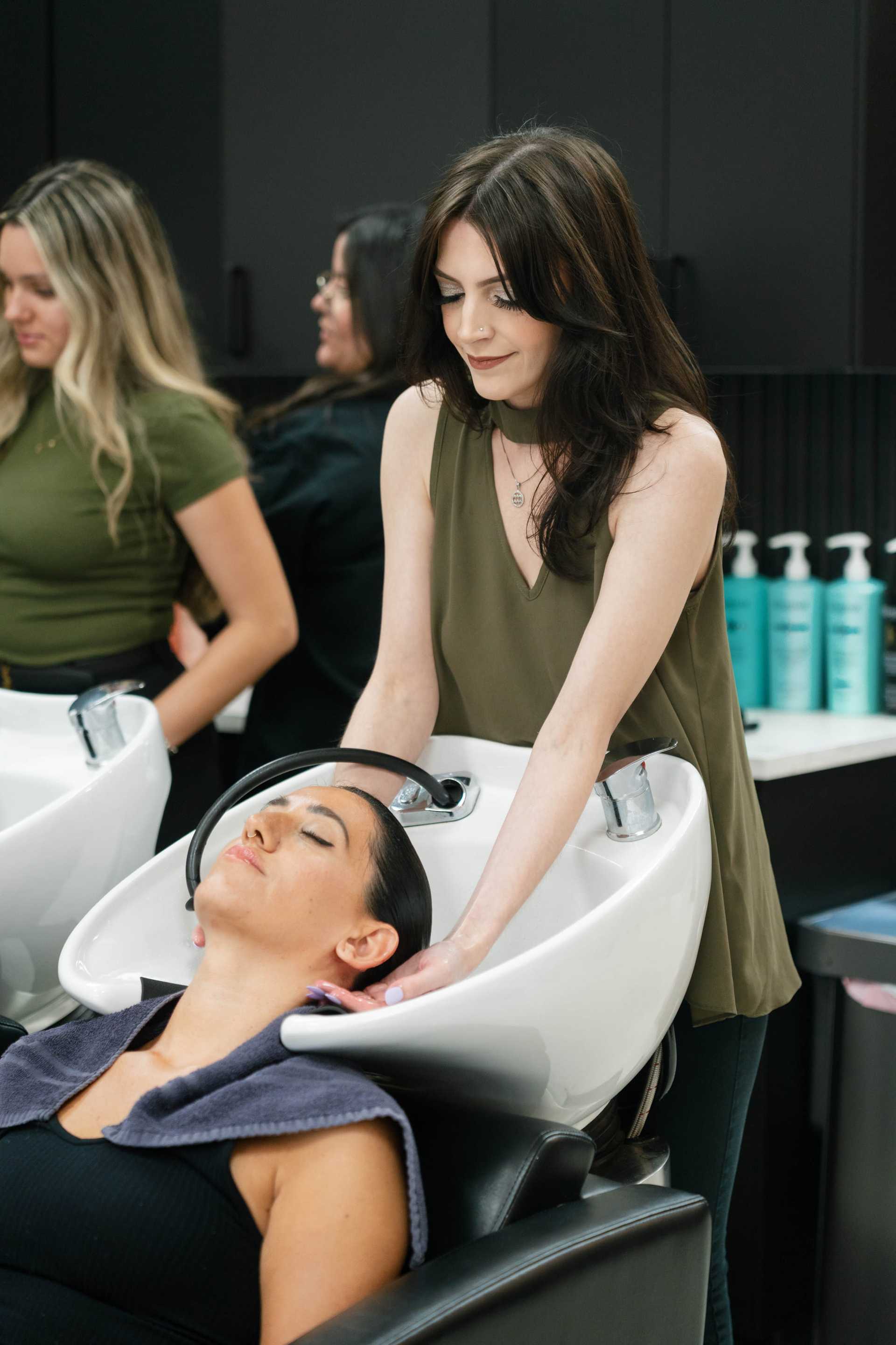 Hair stylist washing woman's hair at a salon sink with two other stylists in the background.