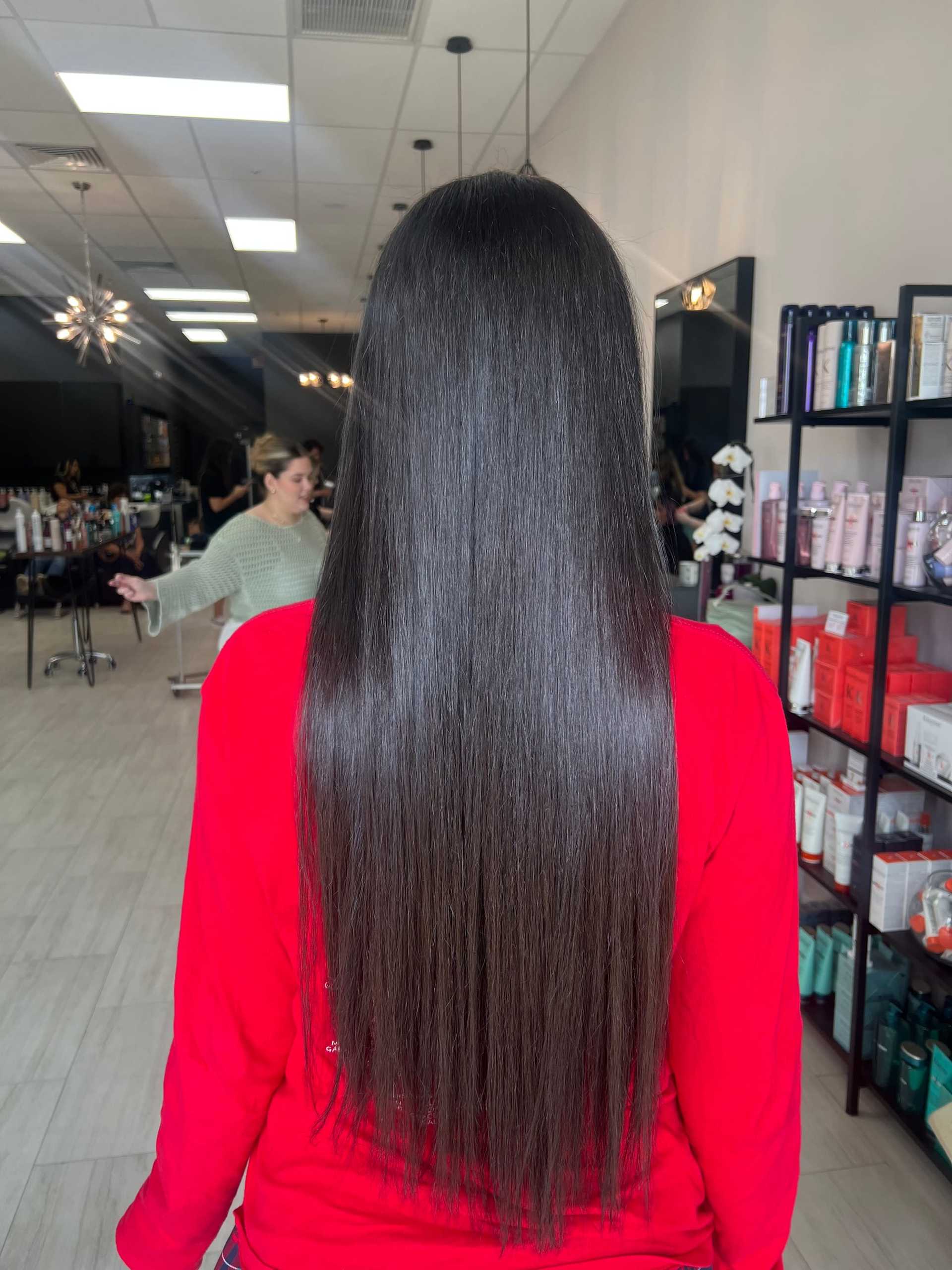 Long, shiny brown hair in salon setting with hairstylist in background.
